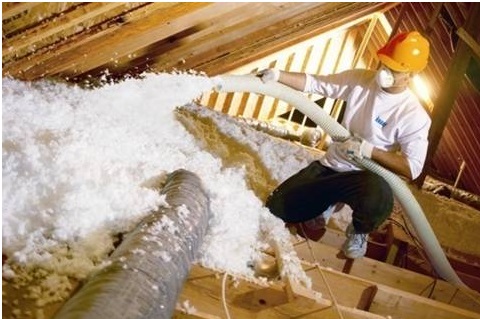 Contractor in a hard hat and face mask installing blown-in insulation in an attic